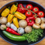 Vue de dessus d'une poêle noire remplie de légumes frais colorés, comprenant des tomates cerises rouges, des champignons, des poivrons jaunes, des piments verts, de l'ail, des oignons et des piments rouges séchés, le tout disposé sur une table en bois.